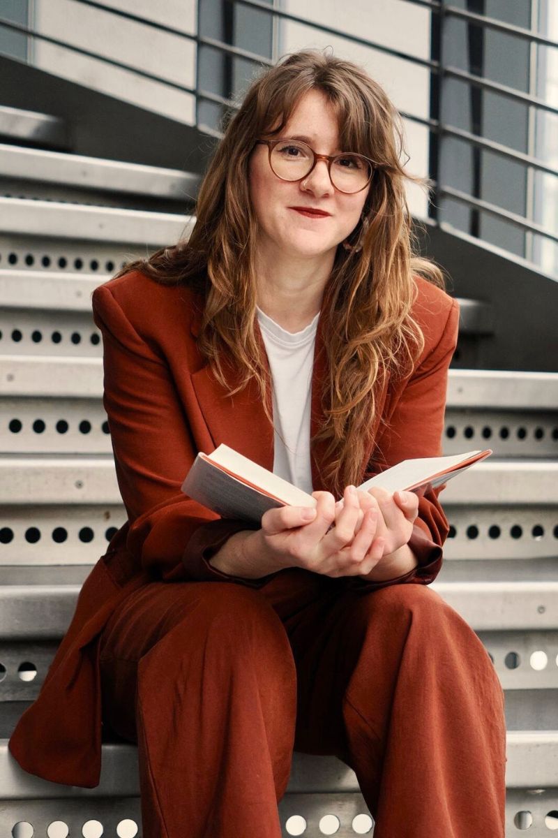 Lea Najork in einem roten Anzug. Sie sitzt auf einer Treppe und hat ein Buch in der Hand.