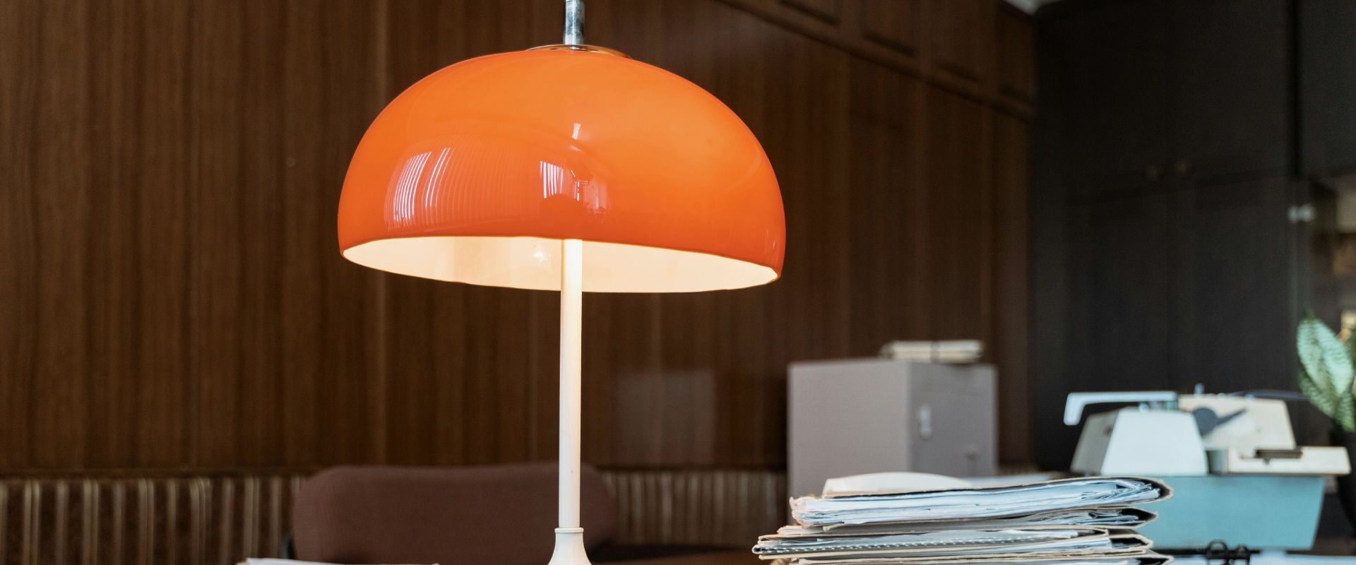 Tischlampe mit rotem Lampenschirm auf einem Schreibtisch, daneben ein Stapel Akten