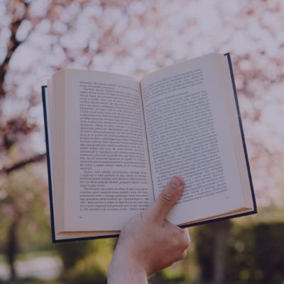 Eine Hand hält ein Buch in die Luft, im Hintergrund sieht man unscharf einen blühenden Baum, es repräsentiert Storytelling.