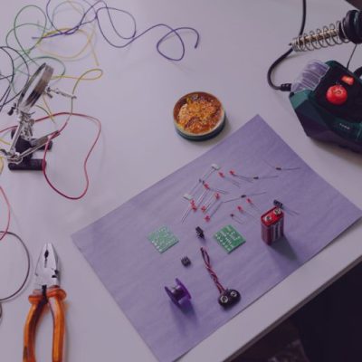 Eine Experimentierstation mit Platinen und Drähten und einer Batterie, daneben ein Lötkölben und eine Zange. Das Bild steht für das Thema Experimentieren und Innovation.